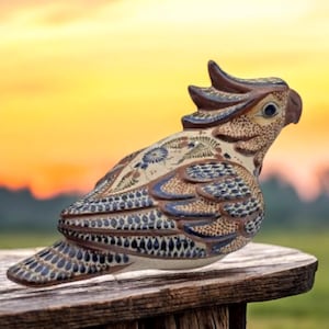 Puede incluir: Figura de pájaro de cerámica con un diseño detallado. El pájaro presenta una cresta, pico y alas estampadas en tonos marrón, azul y crema. La figura está colocada sobre una superficie de madera contra un fondo borroso de puesta de sol.