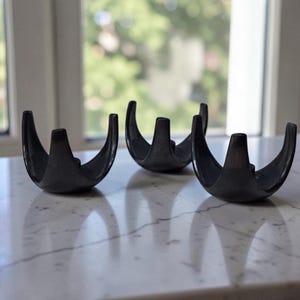 May include: Three dark gray metal candle holders. Each holder has a curved, bowl-like base with three upright prongs. The holders are arranged on a white marble surface, with a window in the background.