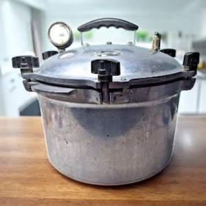 May include: A silver-toned metal pressure canner featuring a circular pressure gauge on the lid, a black plastic handle, and multiple black screw-down clamps around the rim to secure the lid to the main pot body for a tight seal.