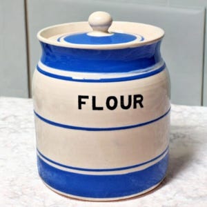 以下が含まれることがあります： 青いストライプと「FLOUR」の文字が入った、クリーム色の陶器製の小麦粉キャニスター。蓋はクリーム色で、青い帯と小さなノブが付いています。キッチン収納に最適です。