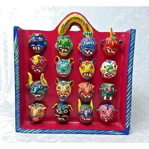 May include: A collection of sixteen colorful, hand-painted, miniature mask sculptures displayed in a red, rectangular wooden frame with a blue border and a yellow handle. Each mask has unique features and expressions.