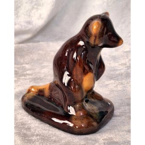 May include: A ceramic bear figurine in shades of brown and tan. The bear is sitting upright on a base, with a glossy finish. The figurine has a detailed design, showcasing the bear's features.