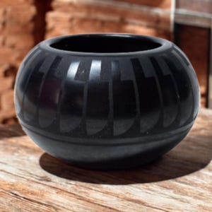 Könnte beinhalten: Eine runde, schwarze Keramikschale mit glänzender Oberfläche. Die Schale hat ein sich wiederholendes geometrisches Muster auf der Außenseite. Die Schale steht auf einer Holzoberfläche.
