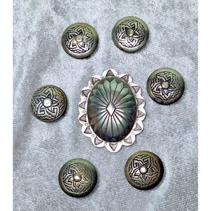 May include: A collection of silver-toned metal embellishments, including six round buttons with a starburst design and an oval-shaped centerpiece. The buttons and centerpiece have an aged, antique appearance, suggesting a vintage or handcrafted origin.