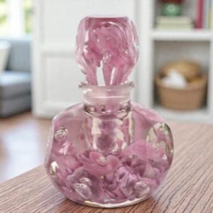 May include: A clear, round glass perfume bottle with a pink tint, filled with pink floral accents. The bottle has a decorative stopper. The bottle is on a wooden surface.
