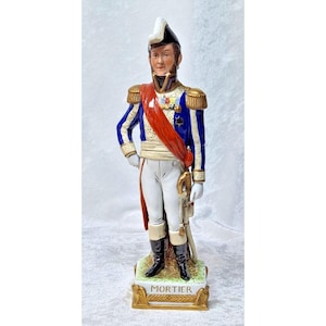 Puede incluir: Figura de porcelana de un oficial militar con uniforme azul y blanco con detalles dorados, una faja roja y un sombrero negro. La figura se encuentra sobre una base con el nombre "MORTIER". La figura sostiene una espada.