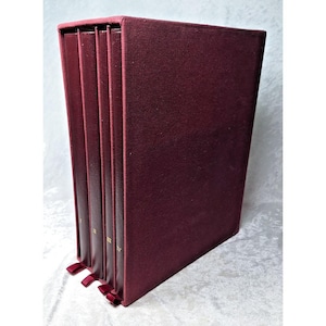 May include: A set of four dark burgundy books housed in a matching slipcase. Each book spine features a gold Roman numeral, labeled I, II, III, and IV, respectively. The set includes small matching burgundy ribbon pulls at the base of each spine.