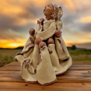 May include: A ceramic sculpture of a figure with outstretched arms, surrounded by smaller figures. The sculpture is primarily beige with brown accents, set against a blurred sunset backdrop. The figures appear to be embracing.