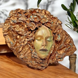 May include: A bronze sculpture of a face with closed eyes, surrounded by textured, crumpled material. The face has a golden hue with green shading. The sculpture is mounted on a wooden surface.