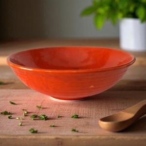 Puede incluir: Un cuenco de cerámica naranja vibrante con un exterior texturizado. El cuenco está sobre una superficie de madera, con una cuchara de madera y hierbas verdes esparcidas cerca. El interior del cuenco es liso y brillante.