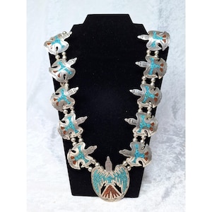 May include: A silver squash blossom necklace with turquoise and coral inlay. The necklace features a central pendant with a bird design and multiple stylized bird-shaped blossoms. The necklace is displayed on a black stand.