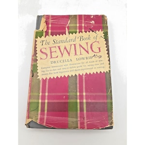 以下が含まれることがあります： Drucella Lowrie著「The Standard Book of Sewing」というタイトルのヴィンテージ裁縫本。赤、緑、黄色のチェック柄の表紙で、タイトルが大きく太字で書かれています。表紙は古く、経年劣化が見られます。