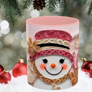 May include: Pink ceramic mug with a snowman design made of paper quilling. The snowman has a pink hat, a brown scarf, and a white face with black eyes and a smile.