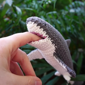 May include: A handmade crochet shark toy featuring a dark gray upper body and a white underbelly. The shark has a small black eye and an open mouth with visible white teeth, crafted with a textured yarn stitch pattern throughout the body.