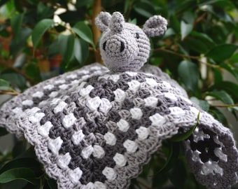 Może przedstawiać: Ręcznie robiony szydełkowy kocyk zabezpieczający z szarą głową hipopotama i kwadratowym kocykiem z wzorem granny stitch w kolorze szarym i białym. Artykuł przeznaczony jest dla niemowląt i małych dzieci.