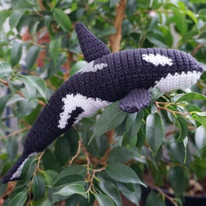 May include: A handmade crochet orca whale toy in black and white, with a dorsal fin and flippers. The plush toy is resting on green leaves, showcasing its detailed craftsmanship and playful design. A charming, handcrafted item.