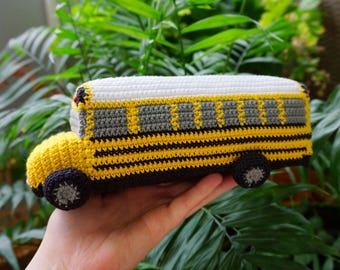 Może przedstawiać: Ręcznie wykonany szydełkowy zabawkowy autobus szkolny z korpusem w kolorze jasnego żółtego, czarnymi poziomymi pasami, białym dachem i szarymi oknami. Autobus posiada ciemnoszare koła z jasnoszarymi środkami oraz zaokrągloną przednią maskę.