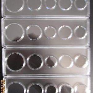 May include: Clear plastic coin holders, each with five circular compartments of varying sizes. The holders are rectangular and stacked, with the coins visible within. The image is well-lit, showing the details of the holders and the coins.
