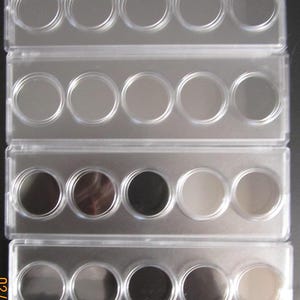 Pode incluir: Porta-moedas de plástico transparente com quatro filas de cinco compartimentos circulares. Alguns compartimentos contêm moedas de cores variadas. O suporte é retangular e transparente.