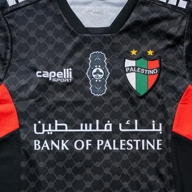 Puede incluir: Camiseta de f&uacute;tbol negra y roja con un logotipo blanco que dice "Capelli Sport" y un escudo con el texto "Palestino" en blanco. La camiseta tambi&eacute;n tiene un patr&oacute;n geom&eacute;trico blanco y negro y el texto "Bank of Palestine" en blanco.