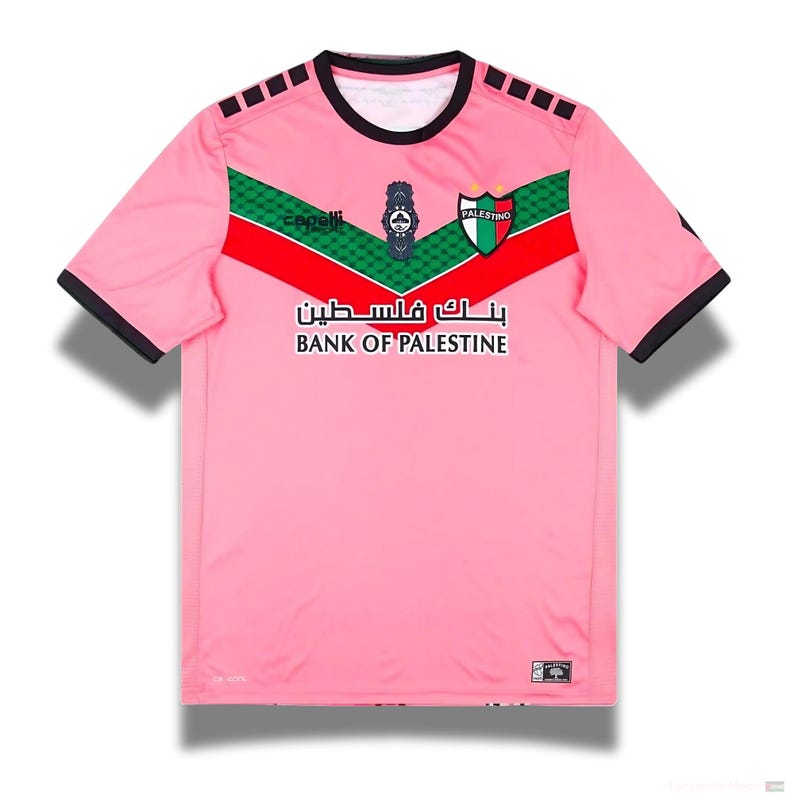 May include: Pink soccer jersey with green and red accents. The jersey has the words "Bank of Palestine" and "بنك فلسطين" on the front. The jersey also has the logo of the Palestinian Football Association.