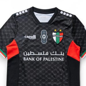 Puede incluir: Camiseta de f&uacute;tbol negra con detalles en verde y rojo. La camiseta tiene un logotipo blanco con el texto "Capelli Sport" y un escudo con el texto "Palestino" y dos estrellas. El texto "Bank of Palestine" est&aacute; impreso en blanco en la parte delantera de la camiseta.