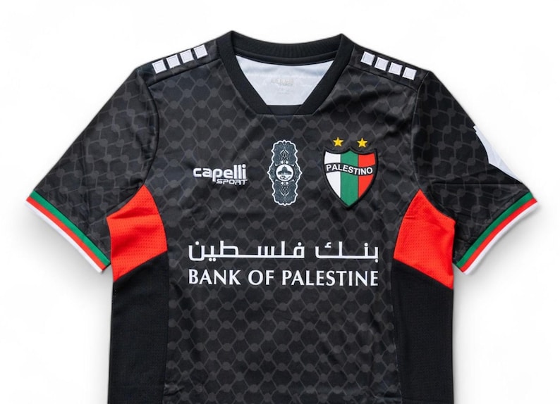 Puede incluir: Camiseta de f&uacute;tbol negra con detalles en verde y rojo. La camiseta tiene un logotipo blanco con el texto "Capelli Sport" y un escudo con el texto "Palestino" y dos estrellas. El texto "Bank of Palestine" est&aacute; impreso en blanco en la parte delantera de la camiseta.