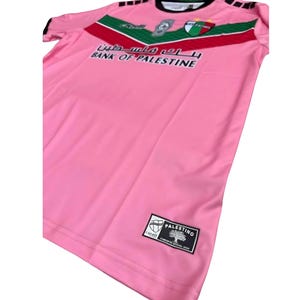 May include: Pink soccer jersey with green and red accents. The jersey has the text "Bank of Palestine" and a logo with an olive tree.