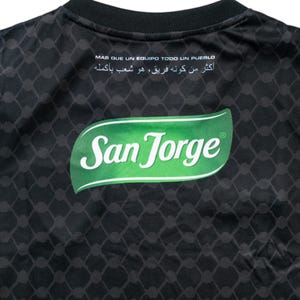 Puede incluir: Camiseta negra con un logotipo "San Jorge" verde y blanco en la espalda. La camiseta tiene un patr&oacute;n geom&eacute;trico en blanco y negro.