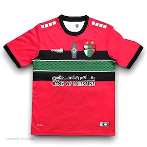 May include: Red football shirt with black and green stripes, featuring the team logo and the words "Bank of Palestine".