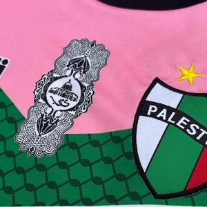 May include: A pink, green, and red soccer jersey with the word "capelli sport" in black lettering on the left side. The jersey also features a white emblem with Arabic script and a green, white, and red shield with the word "Palestino" in white lettering.