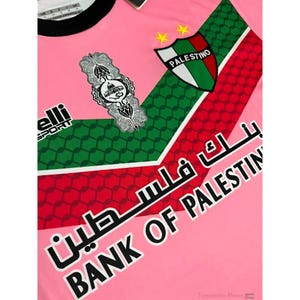 May include: Pink soccer jersey with green and red geometric patterns. The jersey features a white emblem with Arabic script and a shield with the word "PALESTINO" in white text. The text "BANK OF PALESTINE" is printed in black on the bottom right corner of the jersey.