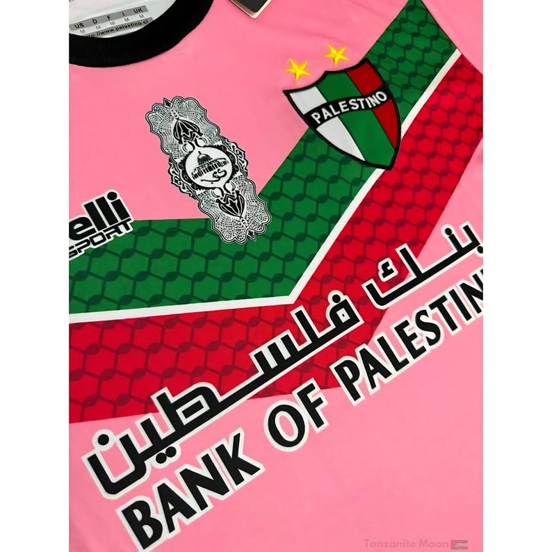 May include: Pink soccer jersey with green and red geometric patterns. The jersey features a white emblem with Arabic script and a shield with the word "PALESTINO" in white text. The text "BANK OF PALESTINE" is printed in black on the bottom right corner of the jersey.