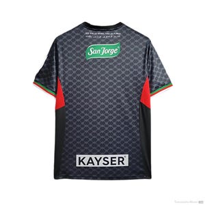 Puede incluir: Camiseta de f&uacute;tbol negra con detalles rojos y verdes. La camiseta tiene un patr&oacute;n de malla negro y gris y presenta el texto "San Jorge" y "KAYSER" en la espalda.
