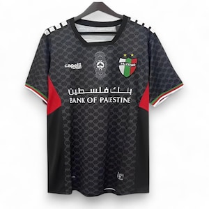 Puede incluir: Camiseta de f&uacute;tbol negra con un logotipo de equipo blanco y verde, las palabras "PALESTINO" y "BANK OF PALESTINE" en blanco, y una franja roja y blanca en el lateral. La camiseta tiene un patr&oacute;n geom&eacute;trico negro y gris.