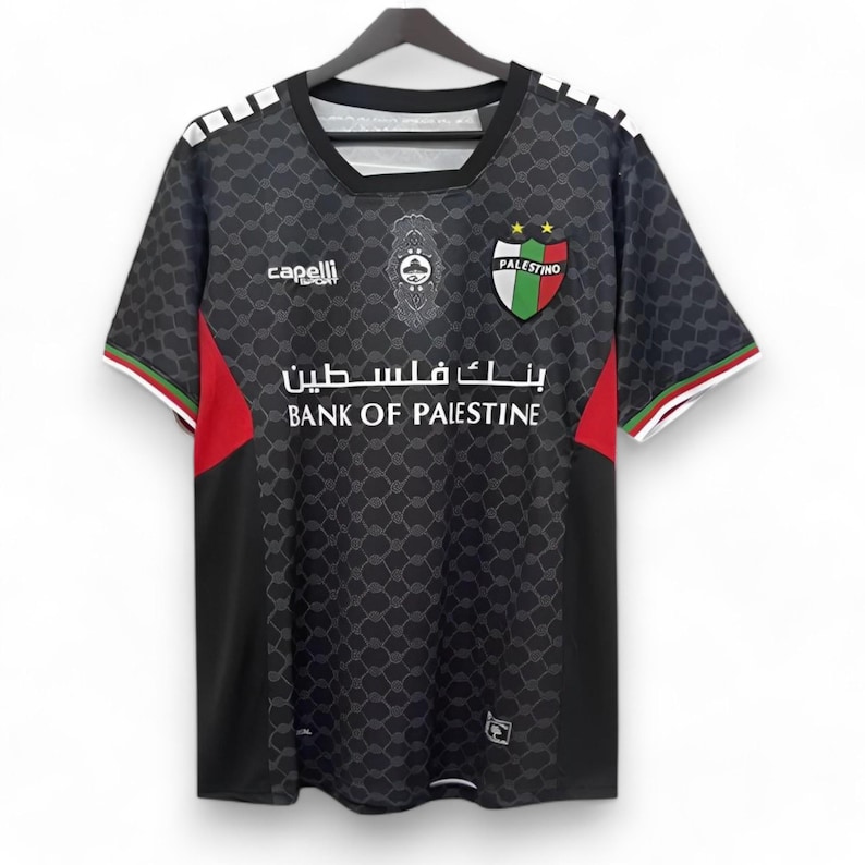 Puede incluir: Camiseta de f&uacute;tbol negra con un logotipo de equipo blanco y verde, las palabras "PALESTINO" y "BANK OF PALESTINE" en blanco, y una franja roja y blanca en el lateral. La camiseta tiene un patr&oacute;n geom&eacute;trico negro y gris.