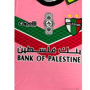 May include: Pink soccer jersey with green and red accents. The jersey features the text "Bank of Palestine" and a logo with two stars and the word "Palestino".