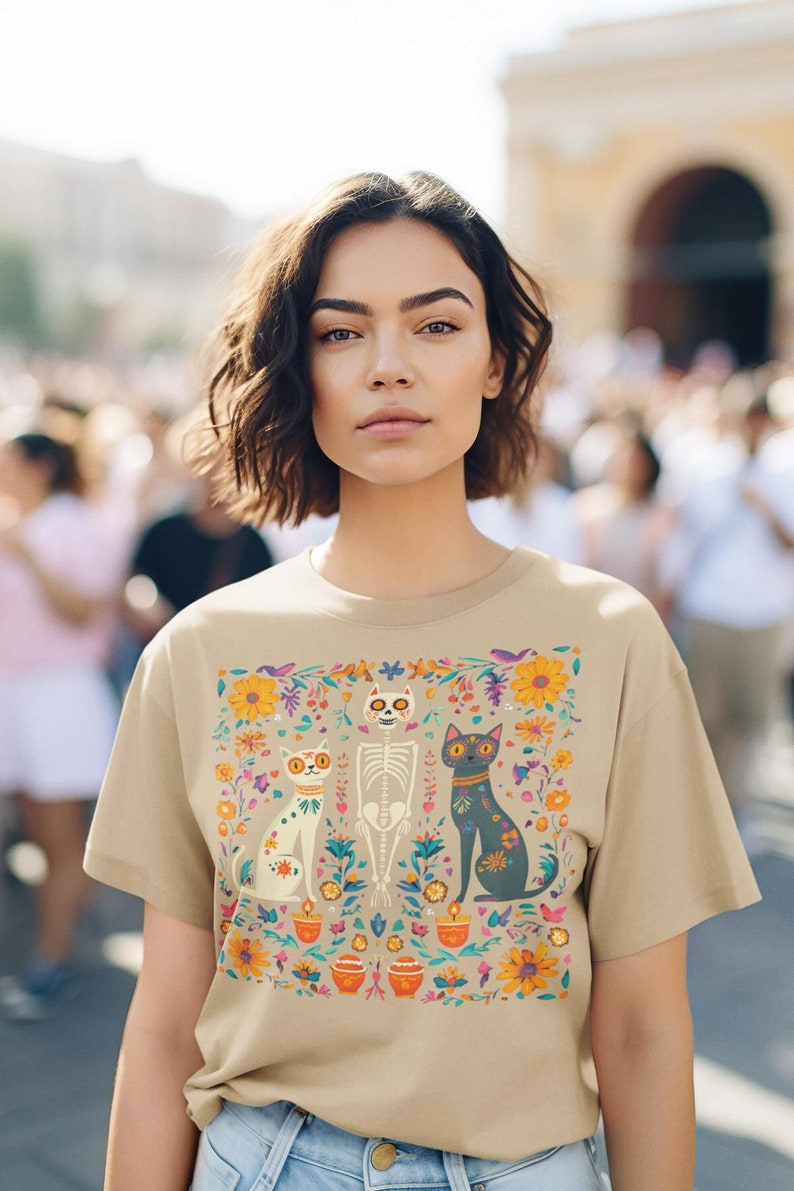 May include: A beige t-shirt with a colorful floral design featuring three cats, one of which is a skeleton. The cats are surrounded by flowers, candles, and other decorative elements.