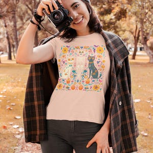 May include: A pink t-shirt with a colorful floral design featuring a skeleton, a cat, and a dog. The design is surrounded by flowers and leaves.