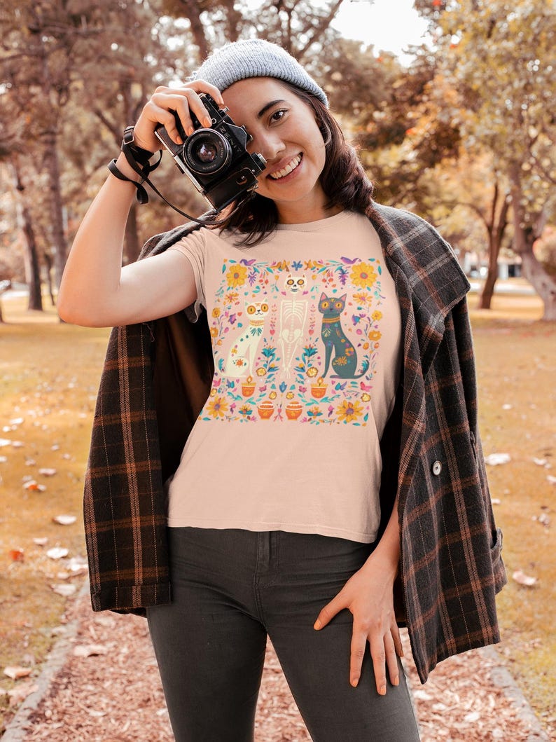 May include: A pink t-shirt with a colorful floral design featuring a skeleton, a cat, and a dog. The design is surrounded by flowers and leaves.