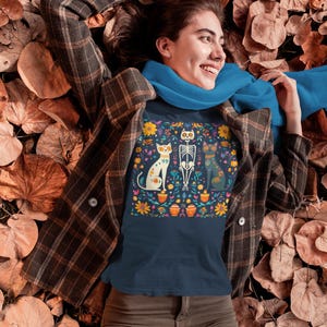 May include: A navy blue t-shirt with a colorful Day of the Dead inspired design featuring three cats and a skeleton. The design includes flowers, skulls, and other traditional Mexican motifs.