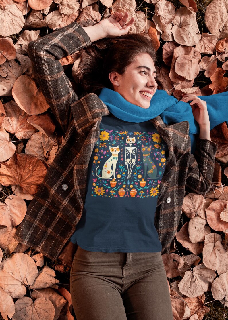 May include: A navy blue t-shirt with a colorful Day of the Dead inspired design featuring three cats and a skeleton. The design includes flowers, skulls, and other traditional Mexican motifs.