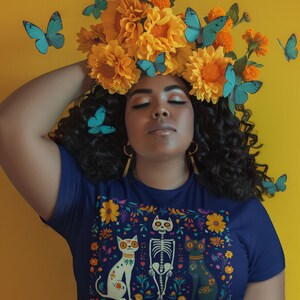 May include: A woman with dark curly hair wears a navy blue t-shirt with a colorful floral design featuring a skeleton and two cats. The design is reminiscent of Day of the Dead imagery. She is wearing a crown of yellow flowers and butterflies.