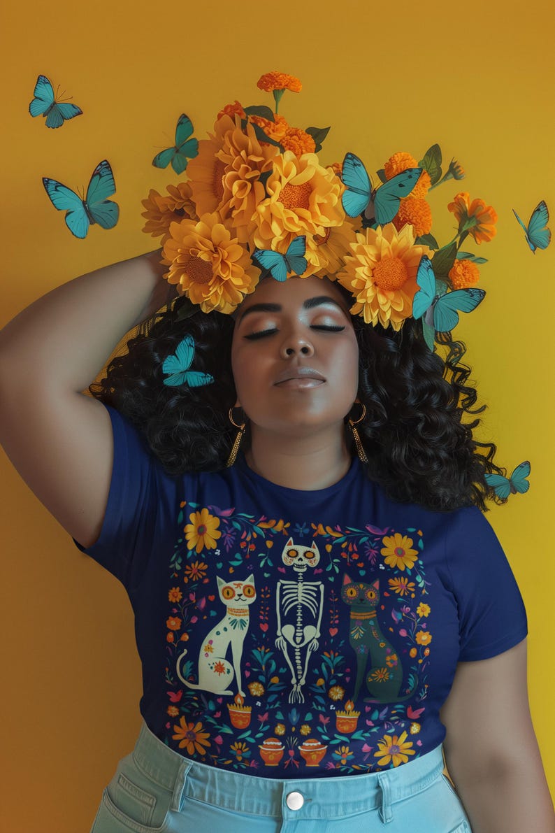 May include: A woman with dark curly hair wears a navy blue t-shirt with a colorful floral design featuring a skeleton and two cats. The design is reminiscent of Day of the Dead imagery. She is wearing a crown of yellow flowers and butterflies.