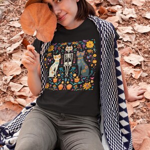 May include: A black t-shirt with a colorful Day of the Dead-inspired design featuring three cats, one of which is a skeleton. The design includes flowers, candles, and other traditional Day of the Dead imagery.