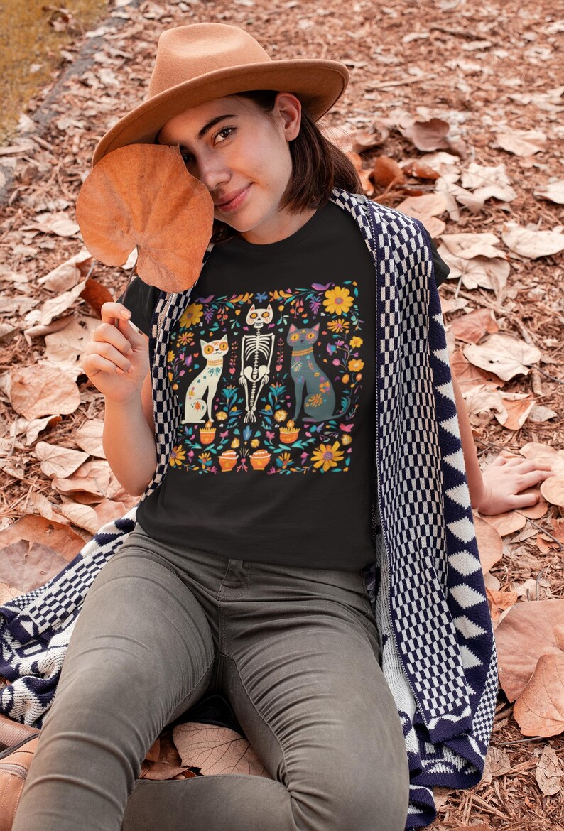 May include: A black t-shirt with a colorful Day of the Dead-inspired design featuring three cats, one of which is a skeleton. The design includes flowers, candles, and other traditional Day of the Dead imagery.