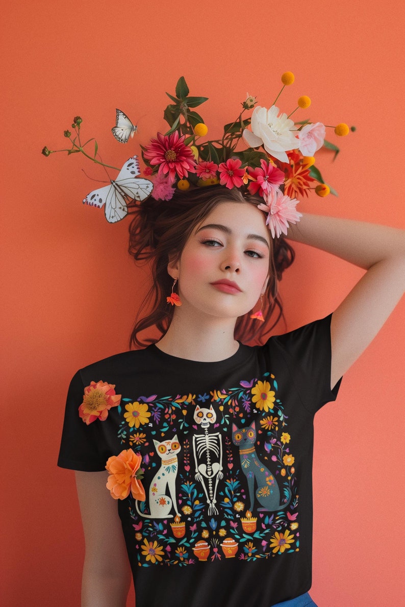 May include: A black t-shirt with a colorful Day of the Dead-inspired design featuring a skeleton and two cats. The design is surrounded by flowers and other decorative elements.