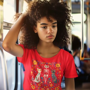 May include: A woman with curly hair is wearing a red t-shirt with a colorful print of three cats and a skeleton. The print features flowers and pots.