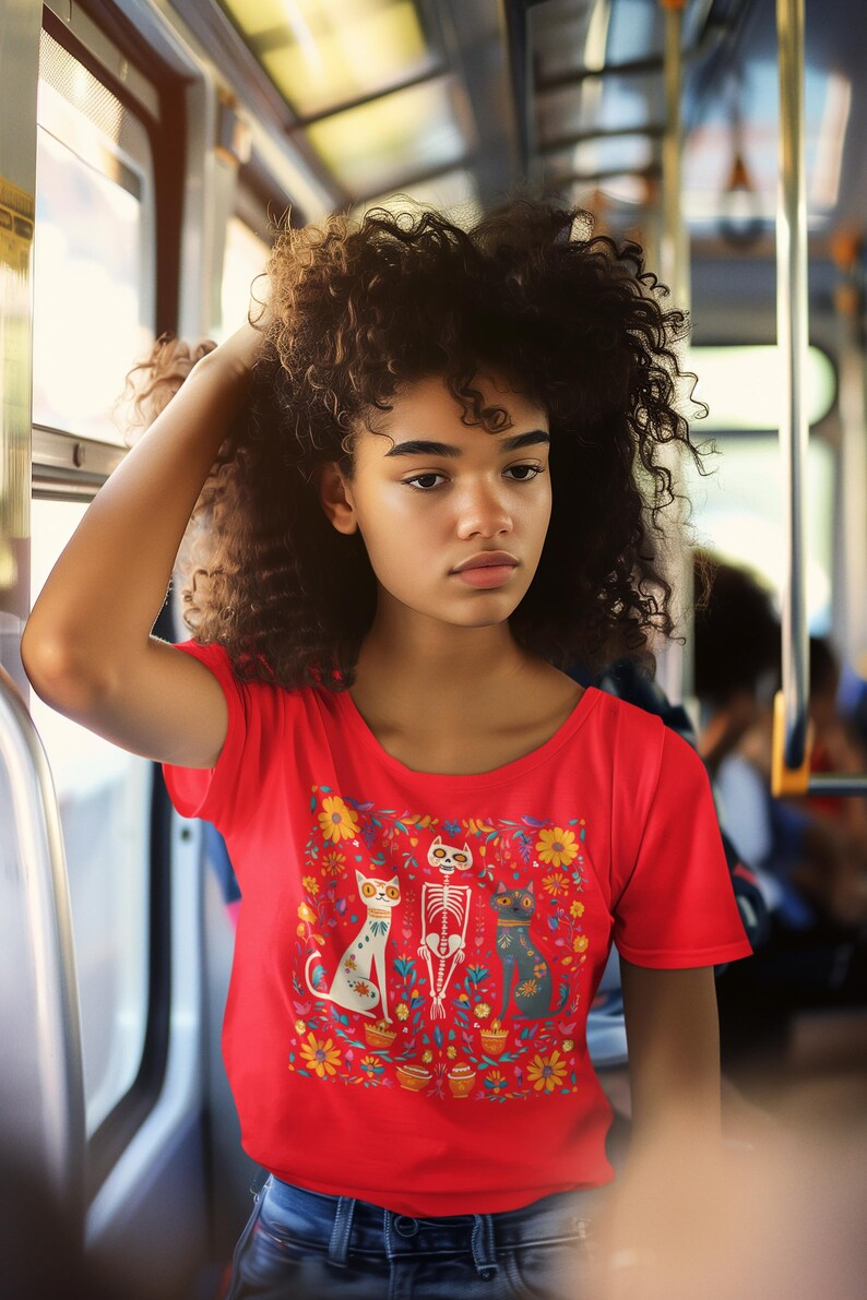 May include: A woman with curly hair is wearing a red t-shirt with a colorful print of three cats and a skeleton. The print features flowers and pots.