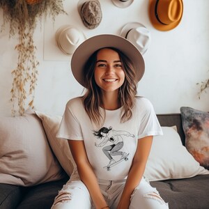 May include: A woman wearing a white t-shirt with a black and white illustration of a person skateboarding. The t-shirt has a crew neck and short sleeves. The woman is smiling and wearing a wide-brimmed hat.